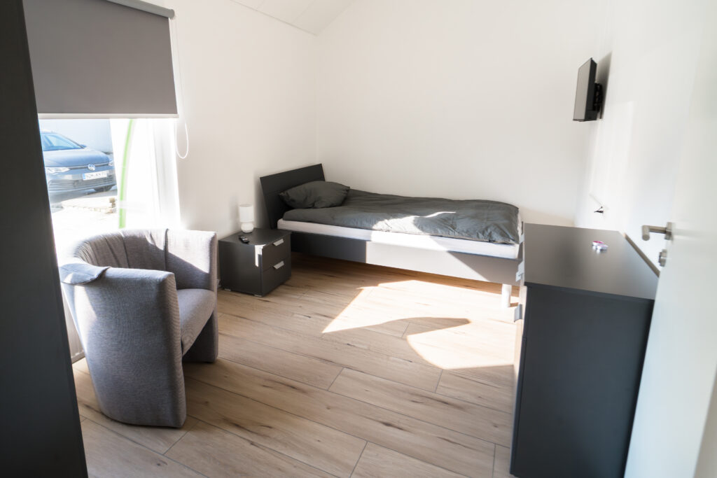 Helles Einzelzimmer mit Bett, grauem Sessel, Nachttisch, Kommode und Wandfernseher.