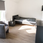 Helles Einzelzimmer mit Bett, grauem Sessel, Nachttisch, Kommode und Wandfernseher.