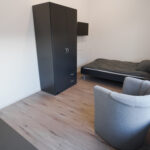 Schlicht eingerichtetes Einzelzimmer mit Bett, Kleiderschrank, Sessel und Fenster.