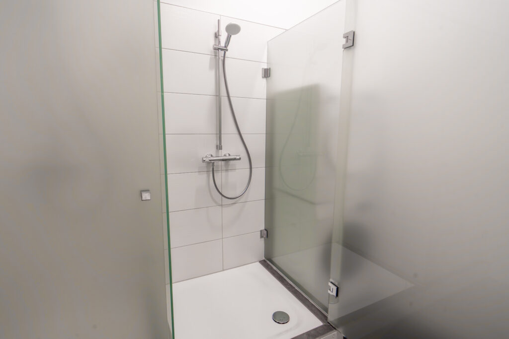 Duschkabine mit Handbrause und Glaswänden in einem weiß gefliesten Badezimmer.