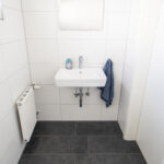 Waschbecken mit Spiegel, Seifenspender und Handtuch in einem schlicht gefliesten Badezimmer.