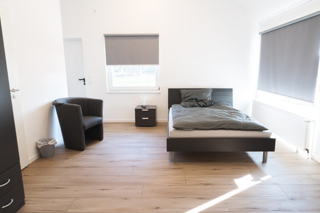 Helles Schlafzimmer mit einem Einzelbett, zwei Fenstern mit Rollos und einem Nachttisch.