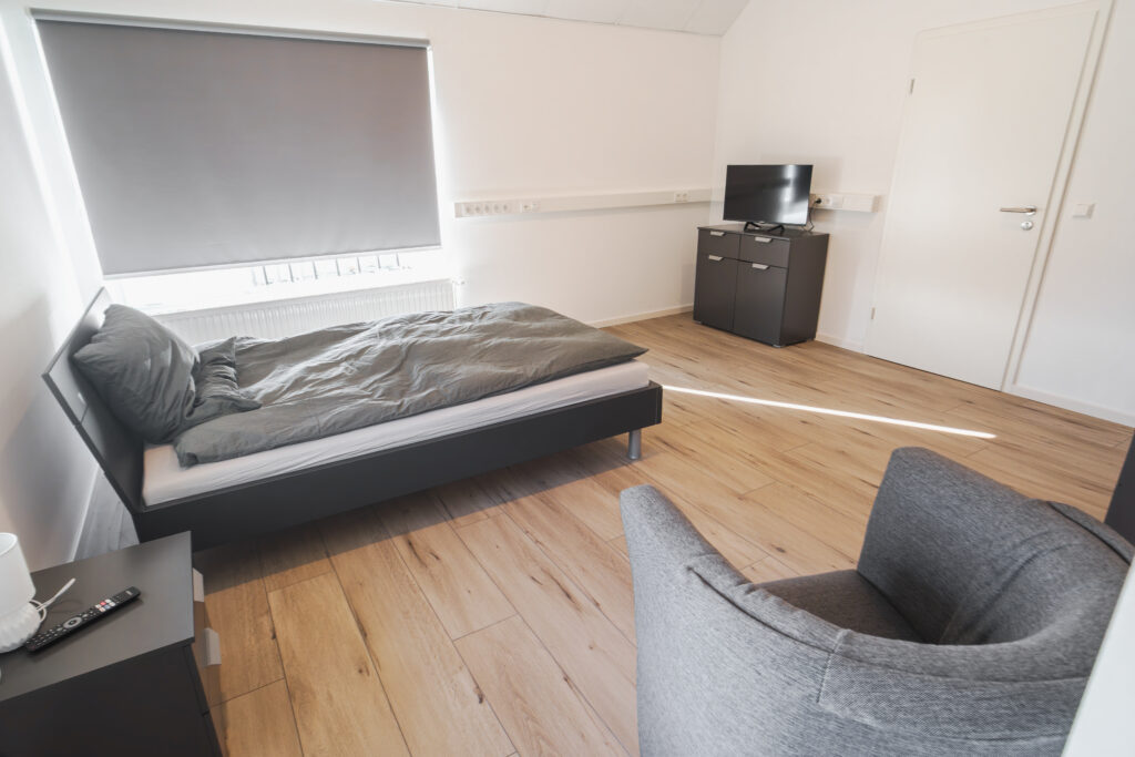 Schlafzimmer mit Einzelbett, Nachttisch, Sessel und Fernseher auf einer Kommode.