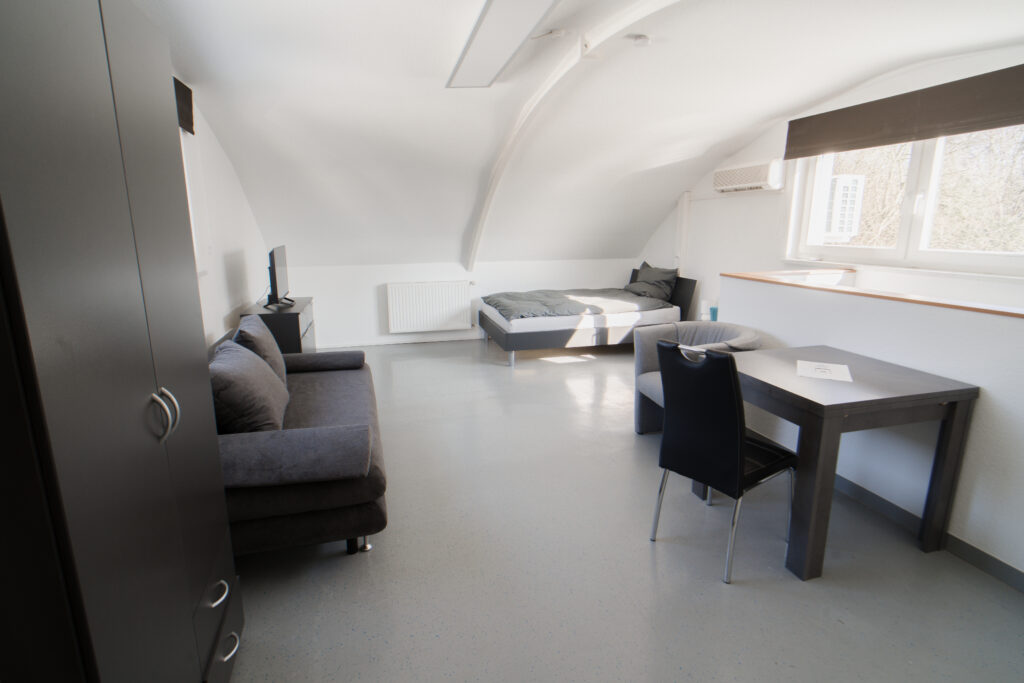 Dachgeschosszimmer mit Bett, Sofa, Tisch und zwei Fenstern mit Verdunkelung.